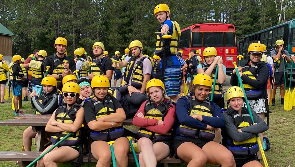 A group of U N E students wearing helmets, life vests, 和 holding paddles